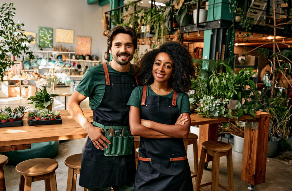 Small flower business. Front view of man andwoman in aprons smiling at camera white standing at own shop.