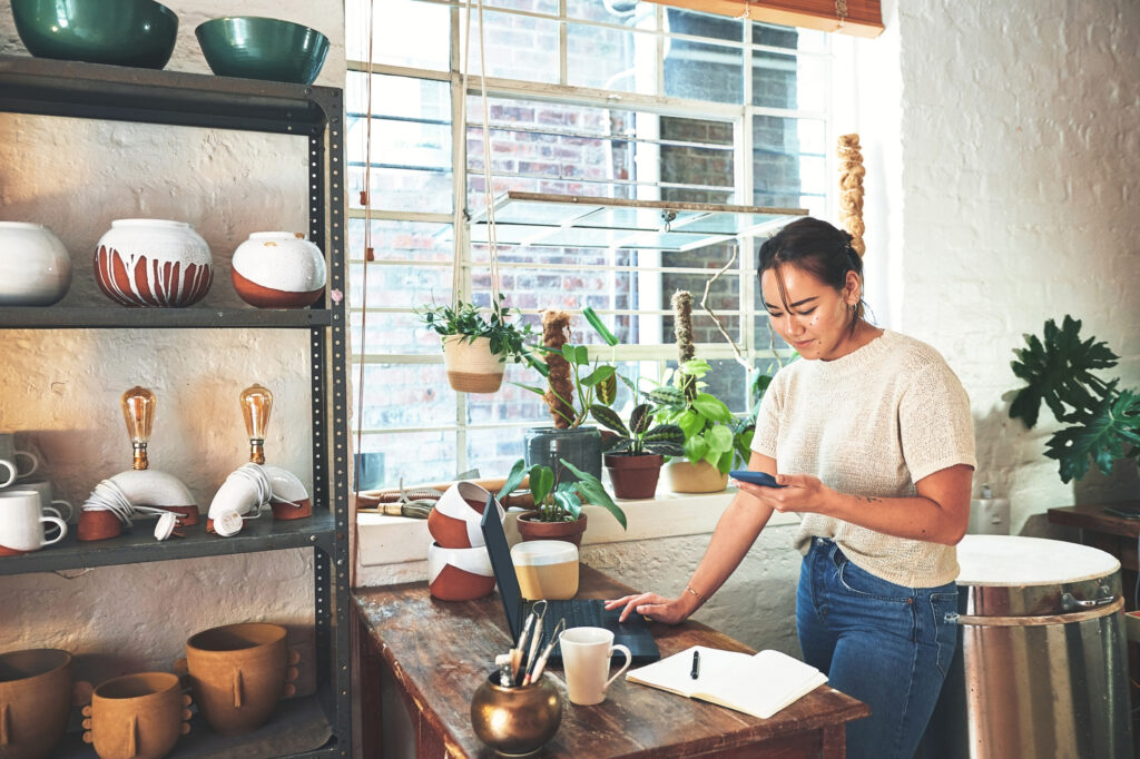 young business owner standing and using her laptop and cellphone in her pottery studio