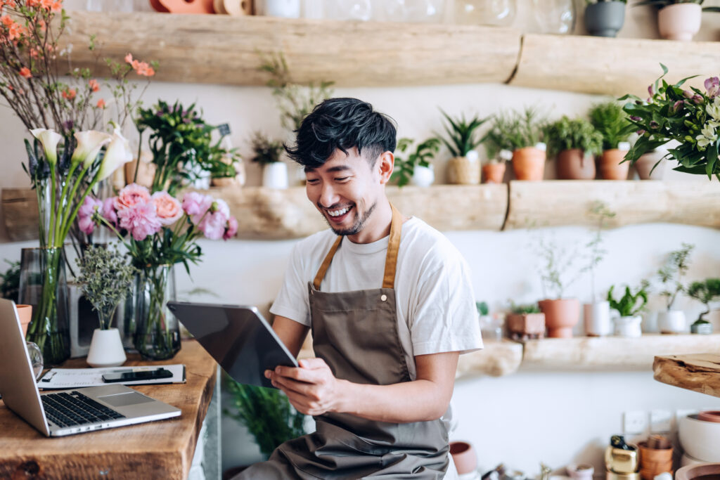 male florist, owner of small business flower shop, using digital tablet while working on laptop